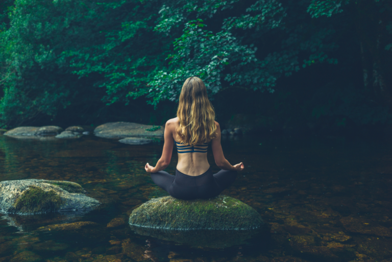 Woman meditating on a rock in a peaceful forest stream, symbolizing mind-body-spirit connection.