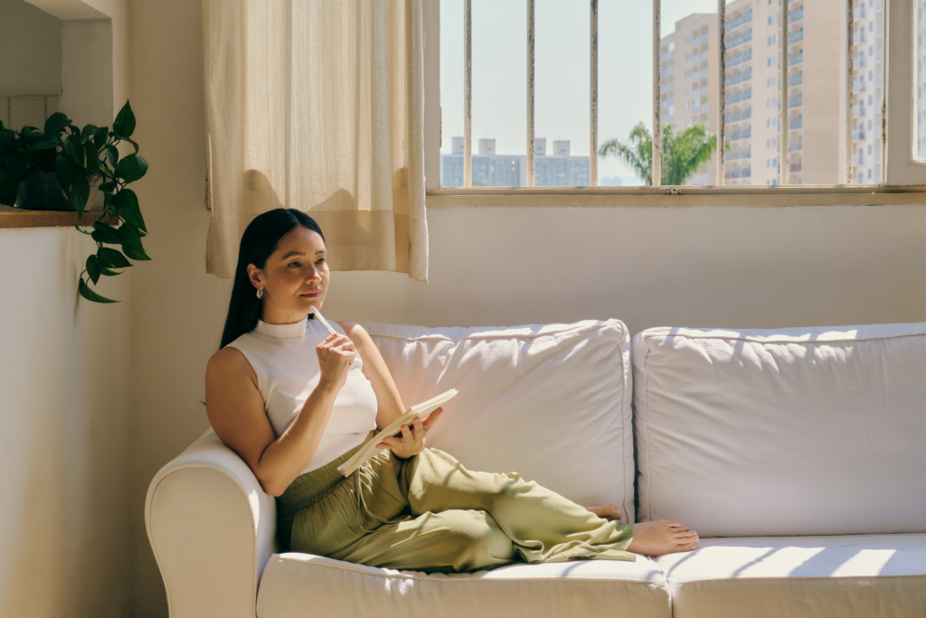A woman sits on a white couch in a sunlit room, holding a notebook and pen, showcasing how journaling helps mental health.